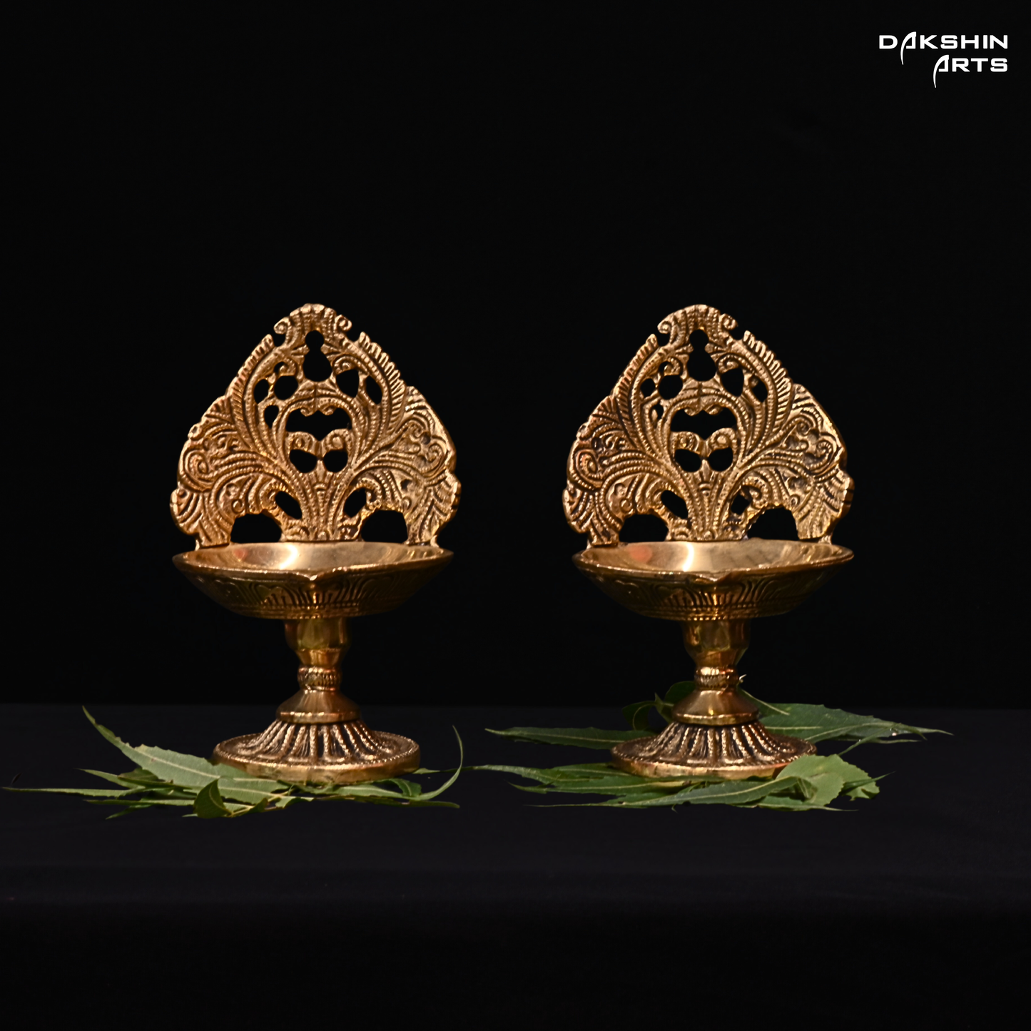 Two ornate brass diya lamps on a black background with 'Dakshin Arts' branding.