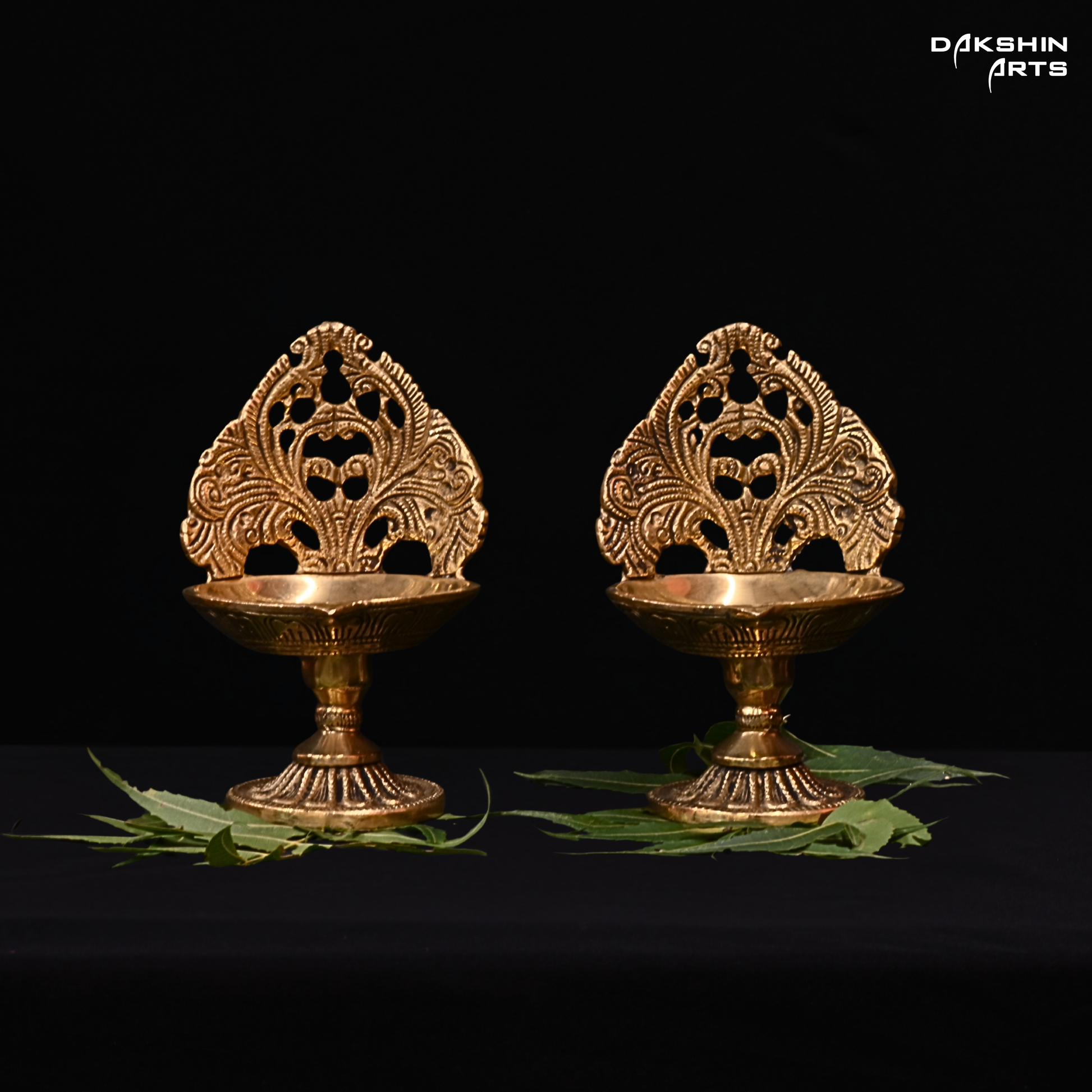 Two ornate brass diya lamps on a black background with 'Dakshin Arts' branding.