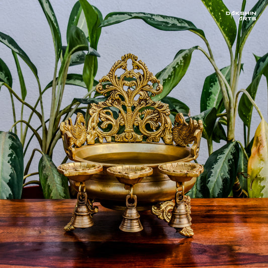 Ornate golden urli with oil lamps with intricate floral and peacock designs, set on a wooden table with leaves in the background- Dakshin Arts