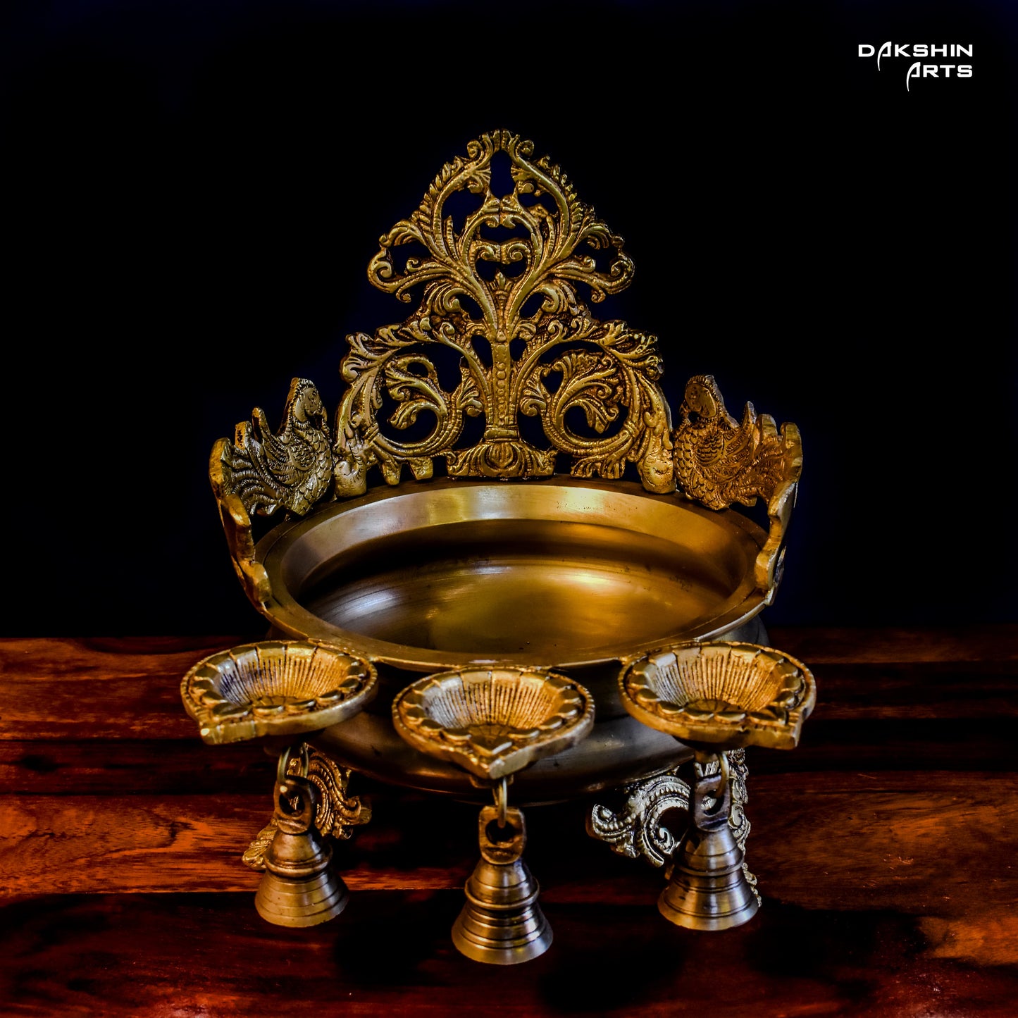 Ornate golden urli with oil lamps with intricate floral and peacock designs, set on a wooden table - Dakshin Arts