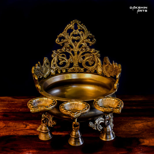 Ornate golden urli with oil lamps with intricate floral and peacock designs, set on a wooden table - Dakshin Arts