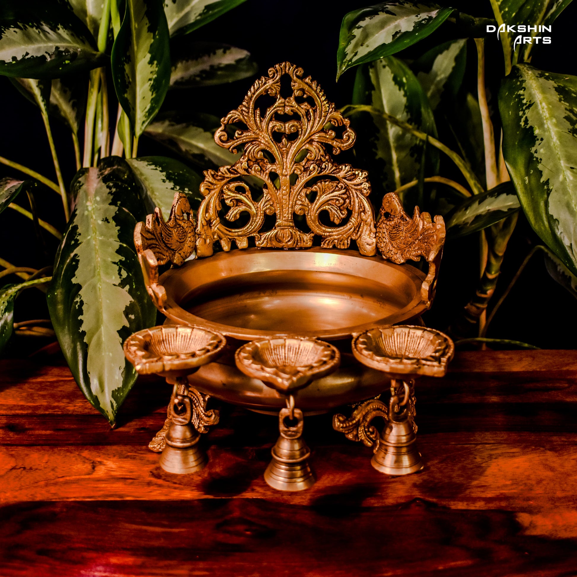 Ornate golden urli with oil lamps with intricate floral and peacock designs, set on a wooden table with leaves in the background- Dakshin Arts