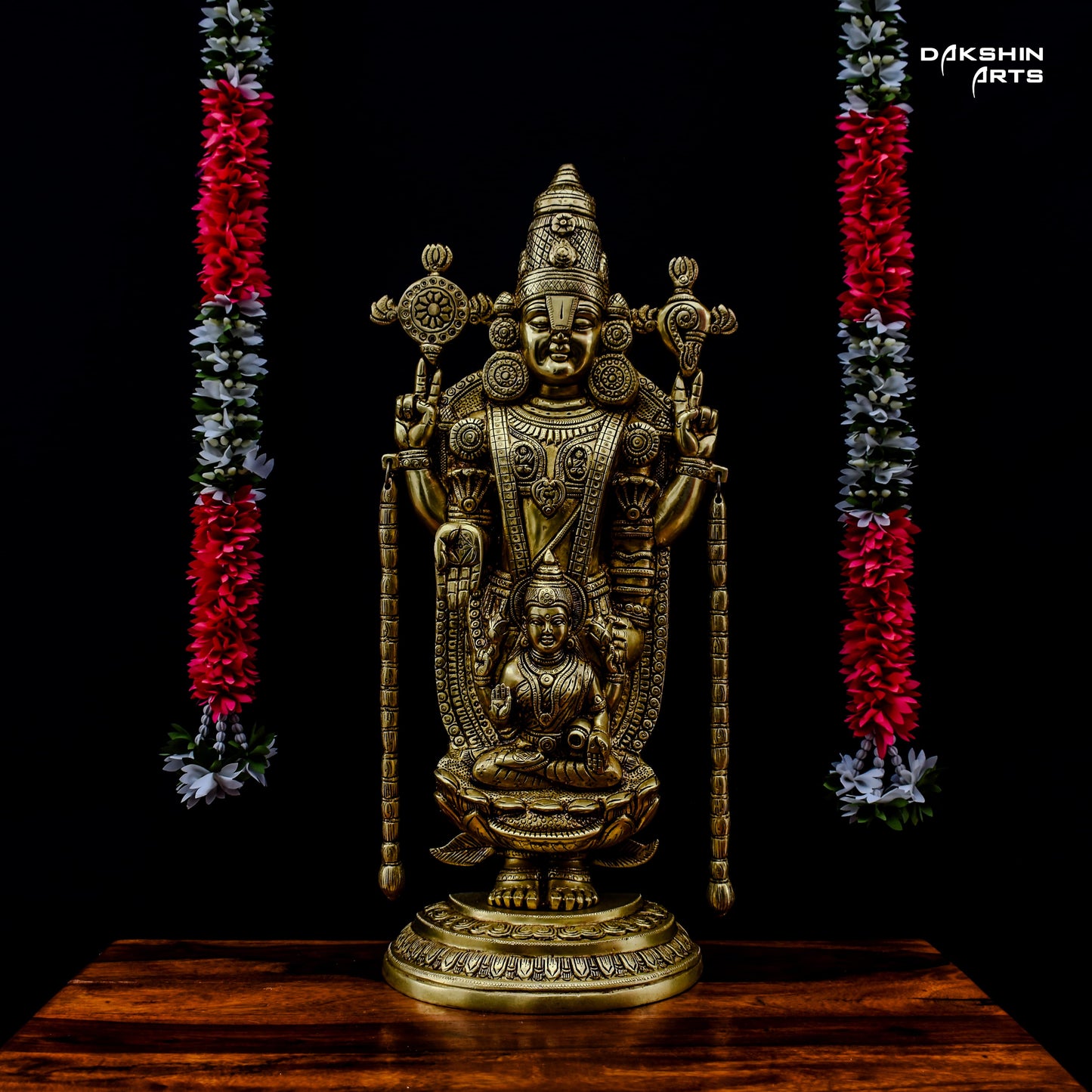 A brass Balaji Padmavati stands on a wooden surface against a black background. It is flanked by flower garlands - Dakshin Arts