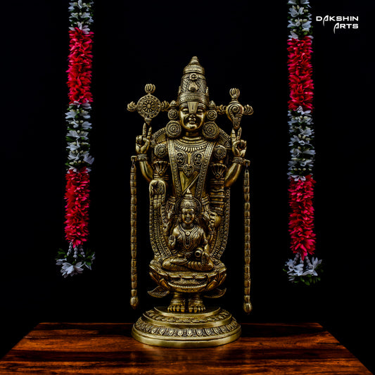 A brass Balaji Padmavati stands on a wooden surface against a black background. It is flanked by flower garlands - Dakshin Arts
