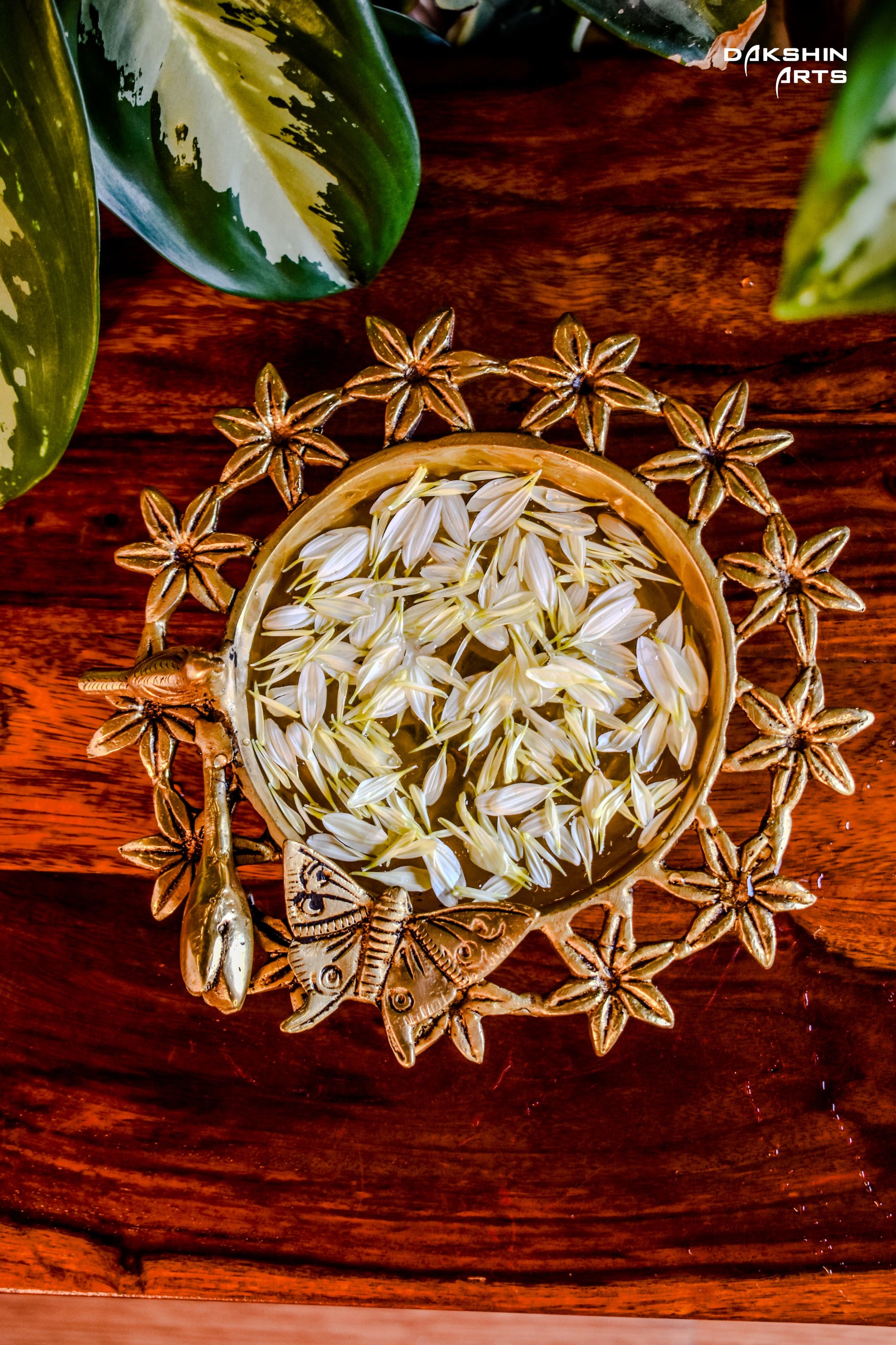 Brass Urli with floral motifs and a butterfly on wooden table filled with white petals. Green leaves peek from the top corner- Dakshin Arts
