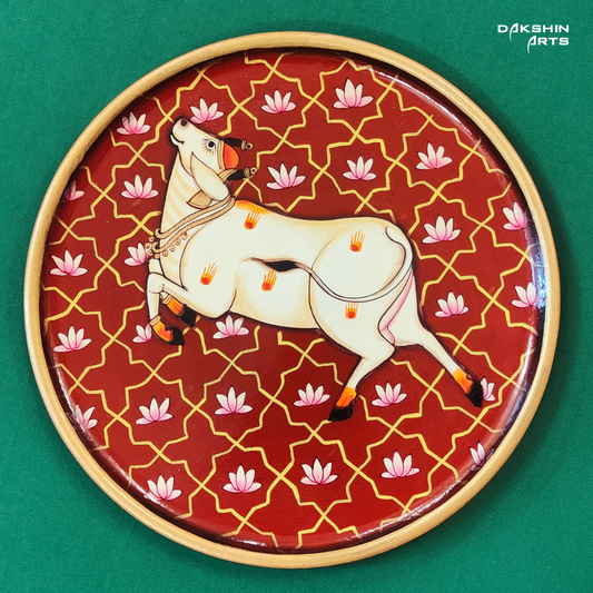 A decorative Pichwai painted wall plate features a stylized white cow set against a red background with pink lotus flowers - Dakshin Arts