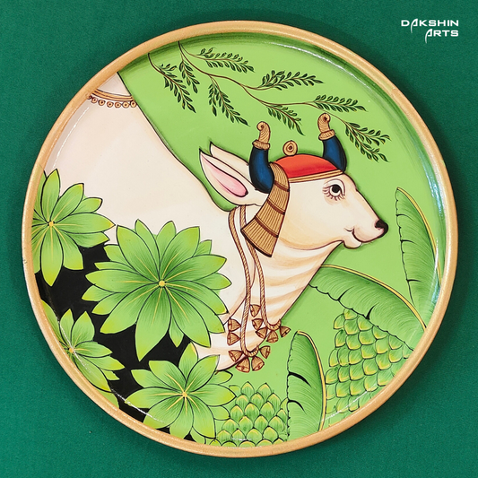 Round Pichwai wall plate with a stylized white cow adorned with gold jewelry on a vibrant green floral background - Dakshin Arts