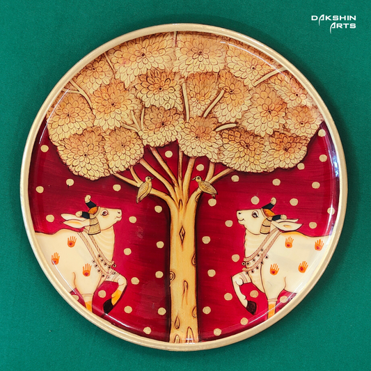 A hand-painted Pichwai wall plate with two white cows adorned with orange handprints, standing under a tree with birds- Dakshin Arts