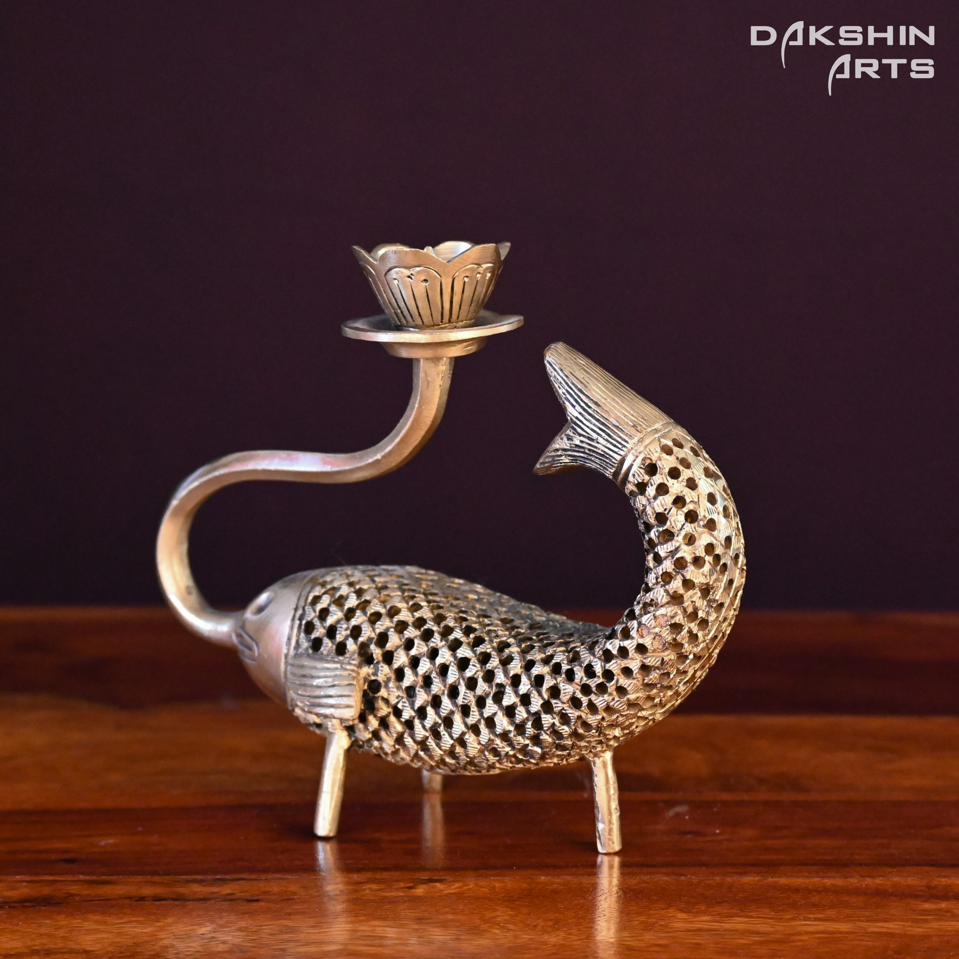 A brass candle holder shaped like a fish with intricate honeycomb detailing and a lotus-shaped top. The fish's mouth curls upward to support the lotus - Dakshin Arts