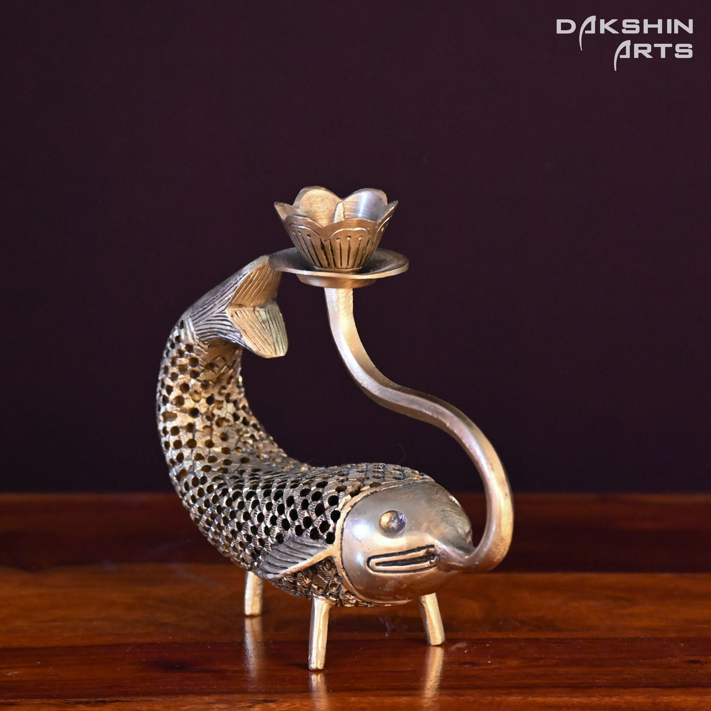 A brass candle holder shaped like a fish with intricate honeycomb detailing and a lotus-shaped top. The fish's mouth curls upward to support the lotus - Dakshin Arts