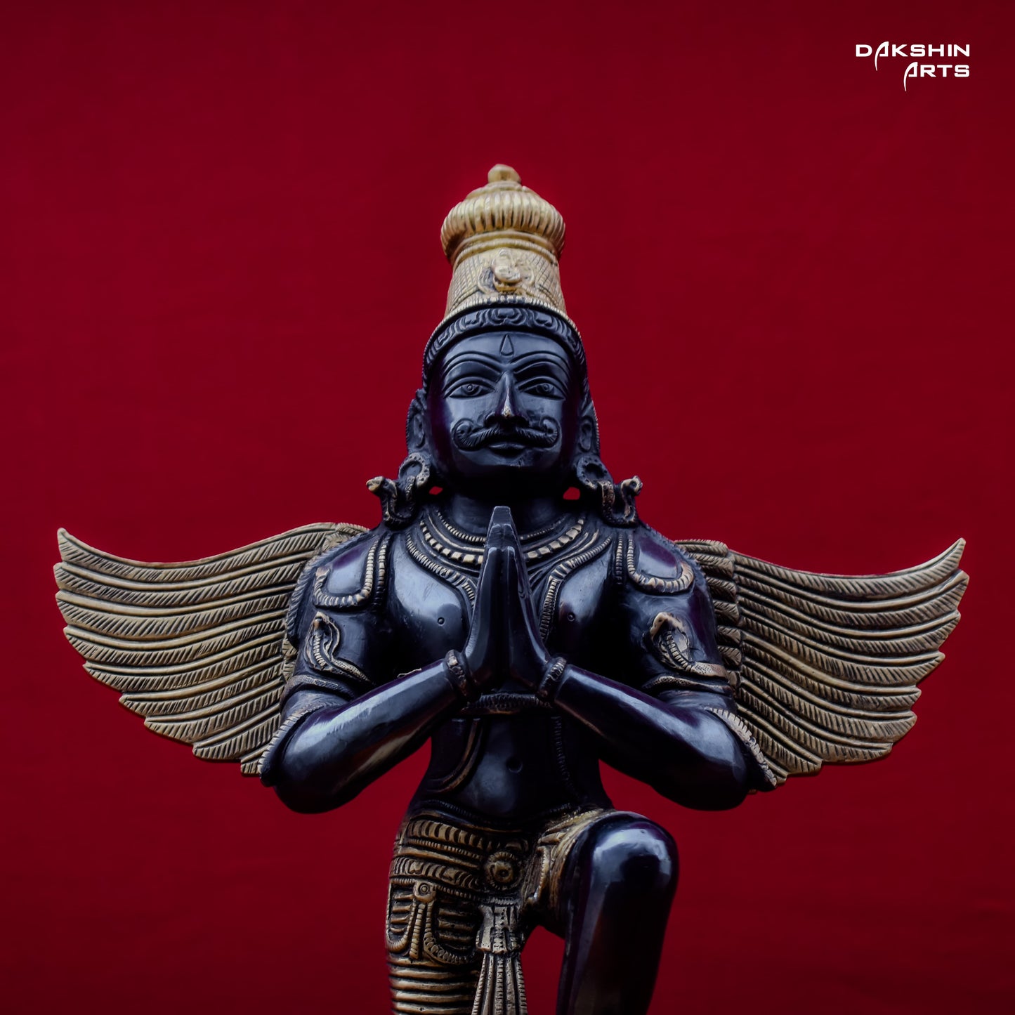 close-up of a black and gold-hued Garuda statue, kneeling, with hands in prayer, set against a rich red background- Dakshin Arts