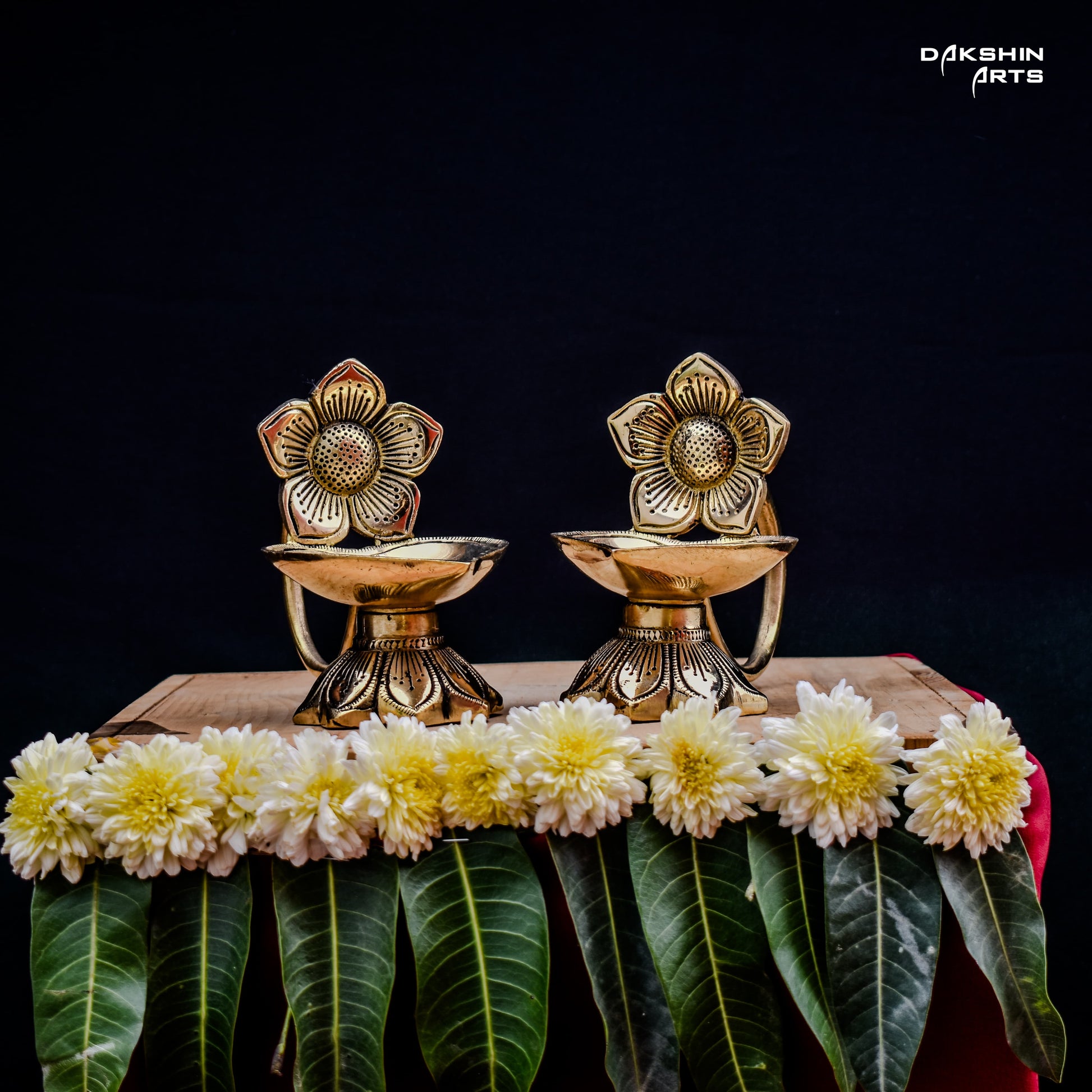 Two ornate brass oil lamps with floral designs sit on a wooden table against a dark background. Elegant and traditional - Dakshin Arts