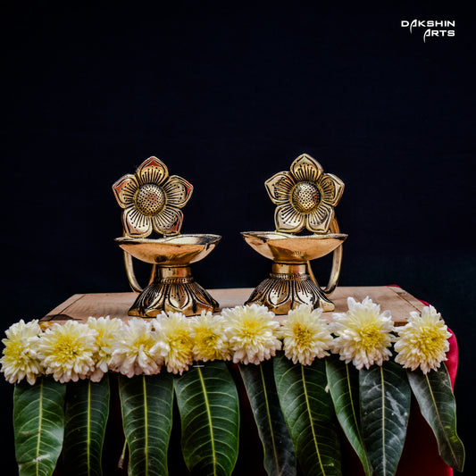 Two ornate brass oil lamps with floral designs sit on a wooden table against a dark background. Elegant and traditional - Dakshin Arts