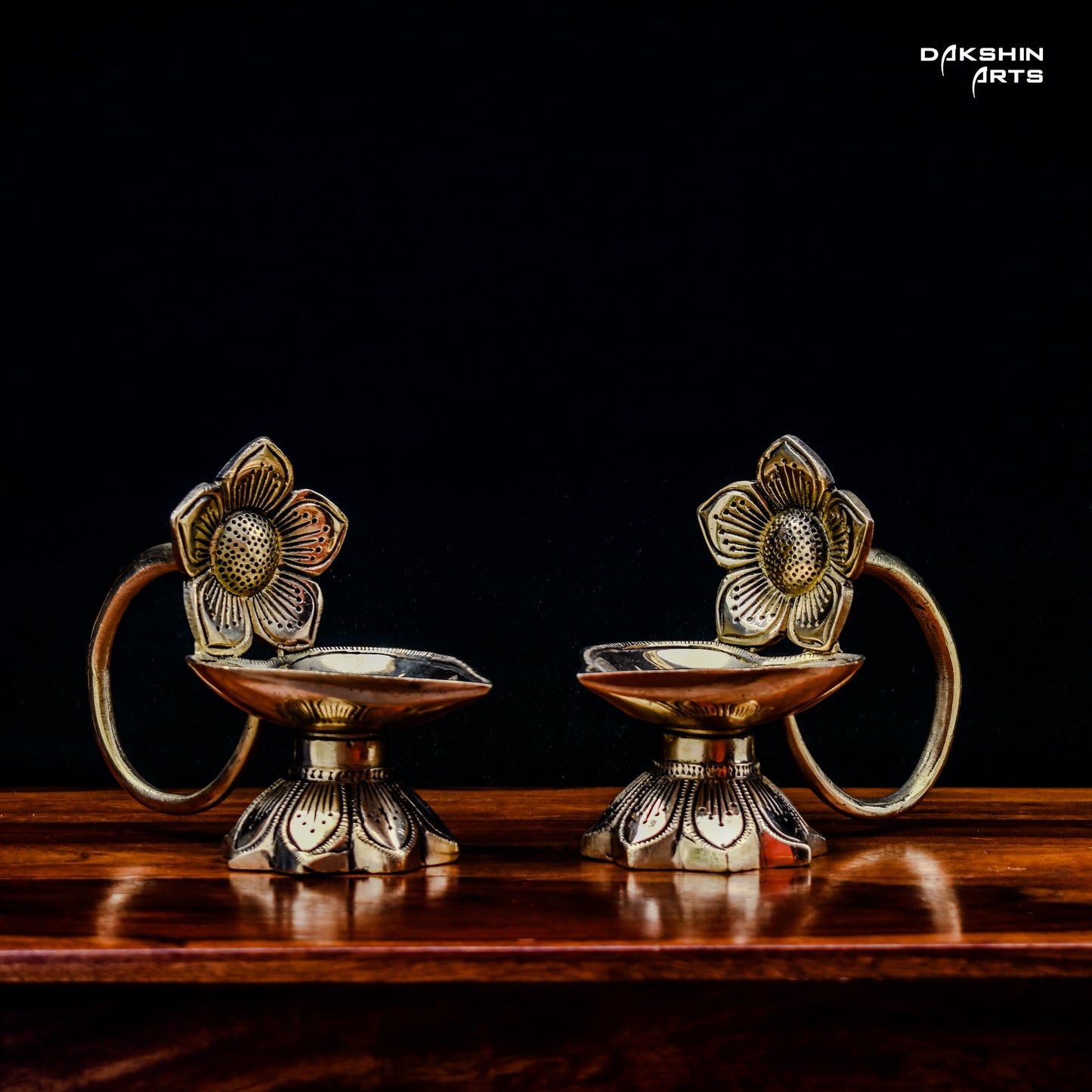 Two ornate brass oil lamps with floral designs sit on a wooden table against a dark background. Elegant and traditional - Dakshin Arts