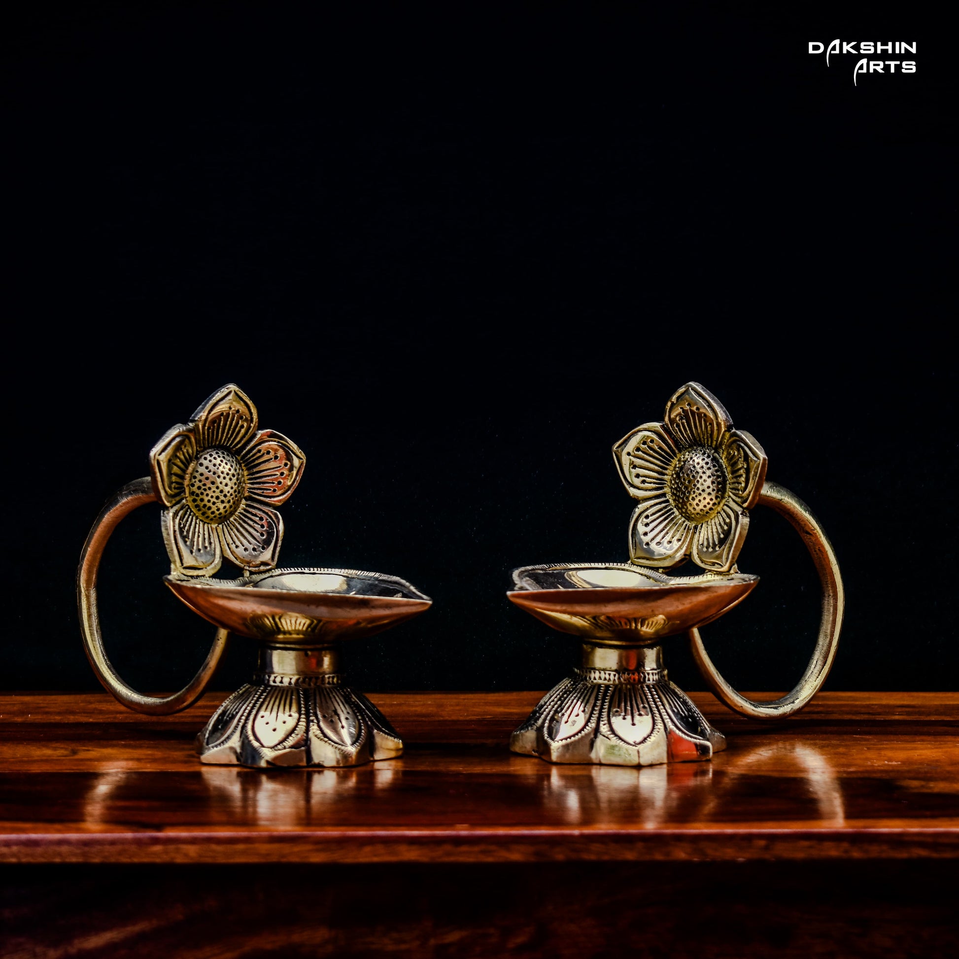 Two ornate brass oil lamps with floral designs sit on a wooden table against a dark background. Elegant and traditional - Dakshin Arts