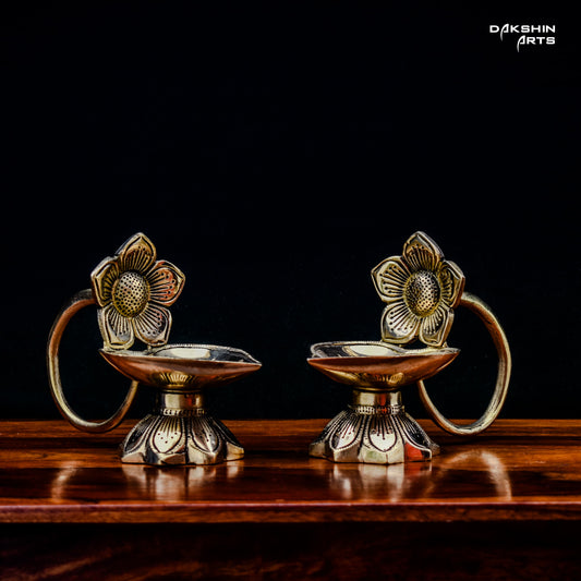 Two ornate brass oil lamps with floral designs sit on a wooden table against a dark background. Elegant and traditional - Dakshin Arts