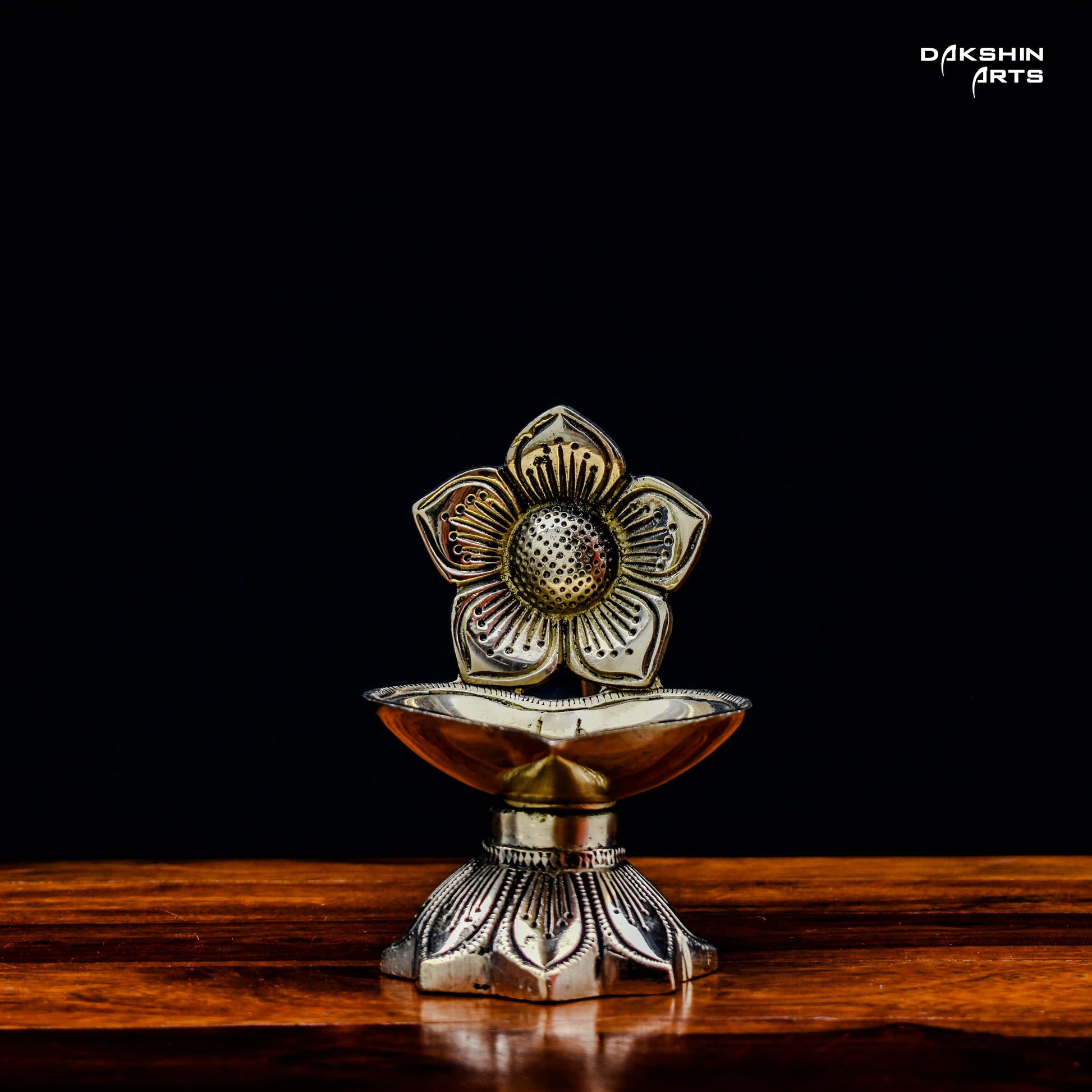 One ornate brass oil lamps with floral designs sit on a wooden table against a dark background. Elegant and traditional - Dakshin Arts