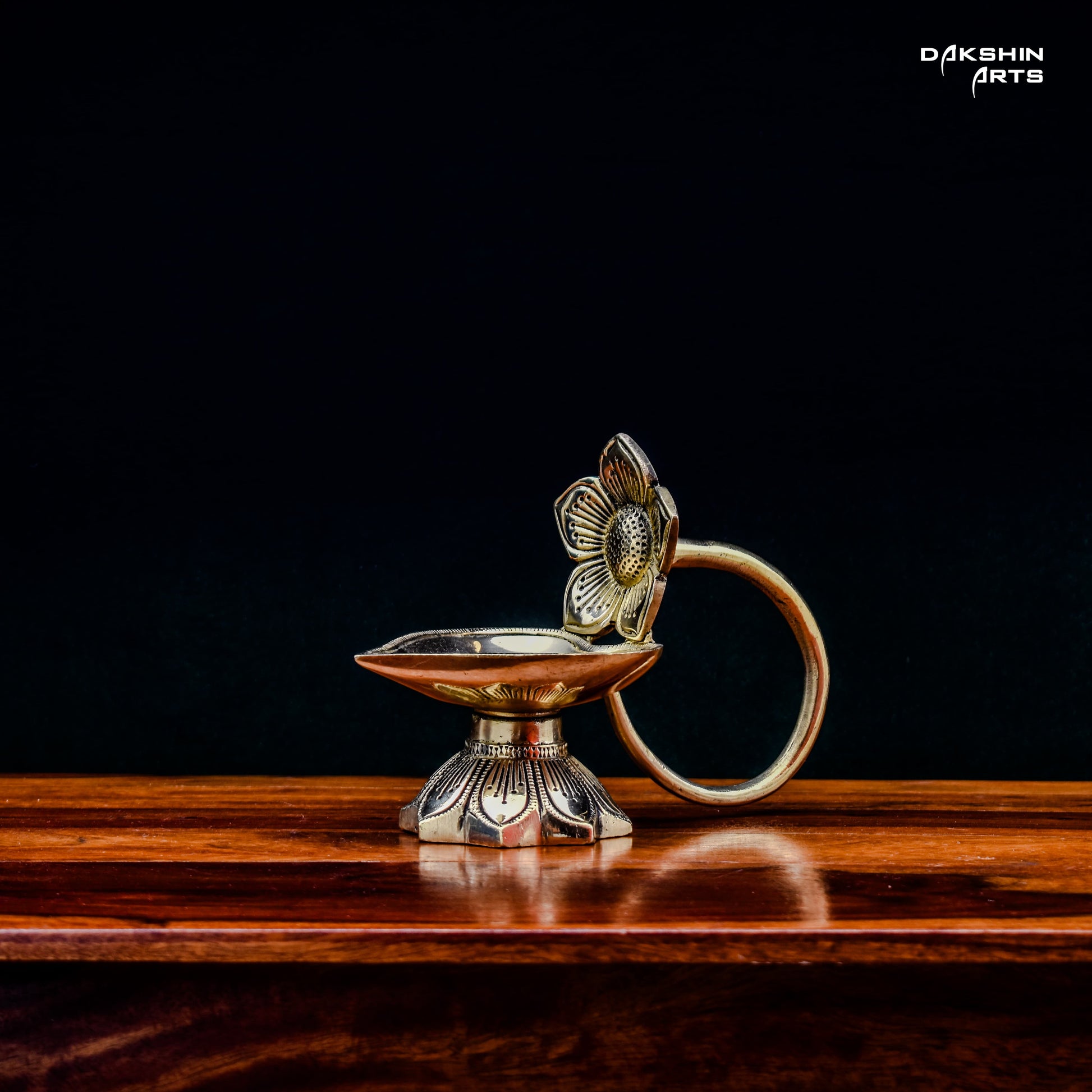 One ornate brass oil lamps with floral designs sit on a wooden table against a dark background. Elegant and traditional - Dakshin Arts