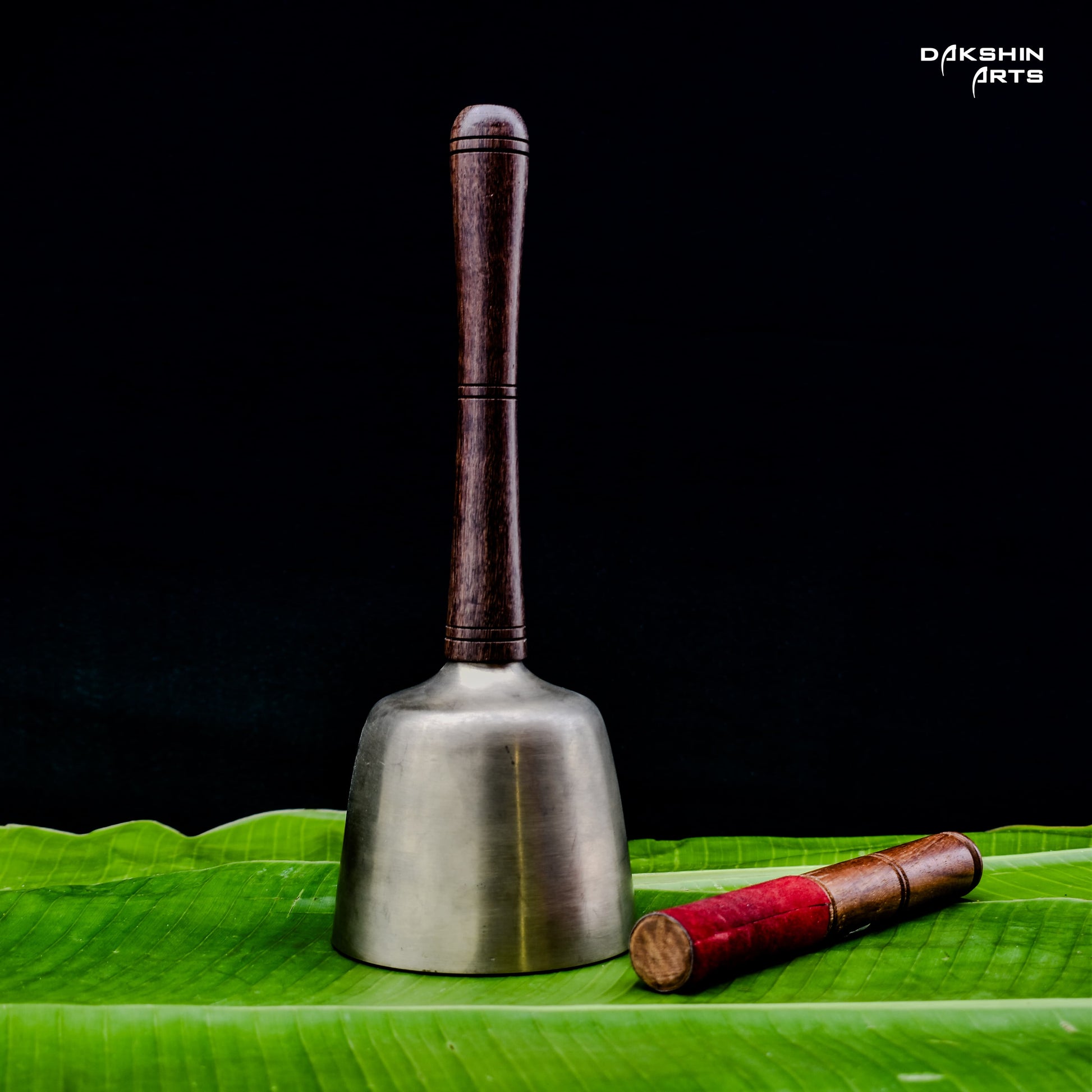 A wooden-handled metal bell stands upright on green banana leaves. Beside it lies a wooden mallet- Dakshin Arts