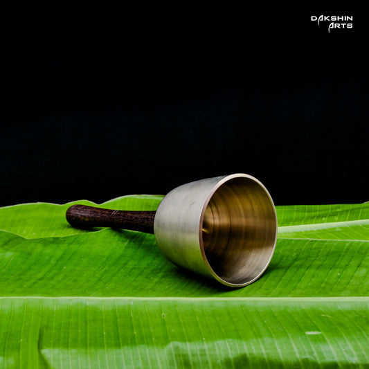 A wooden-handled metal bell stands upright on green banana leaves - Dakshin Arts