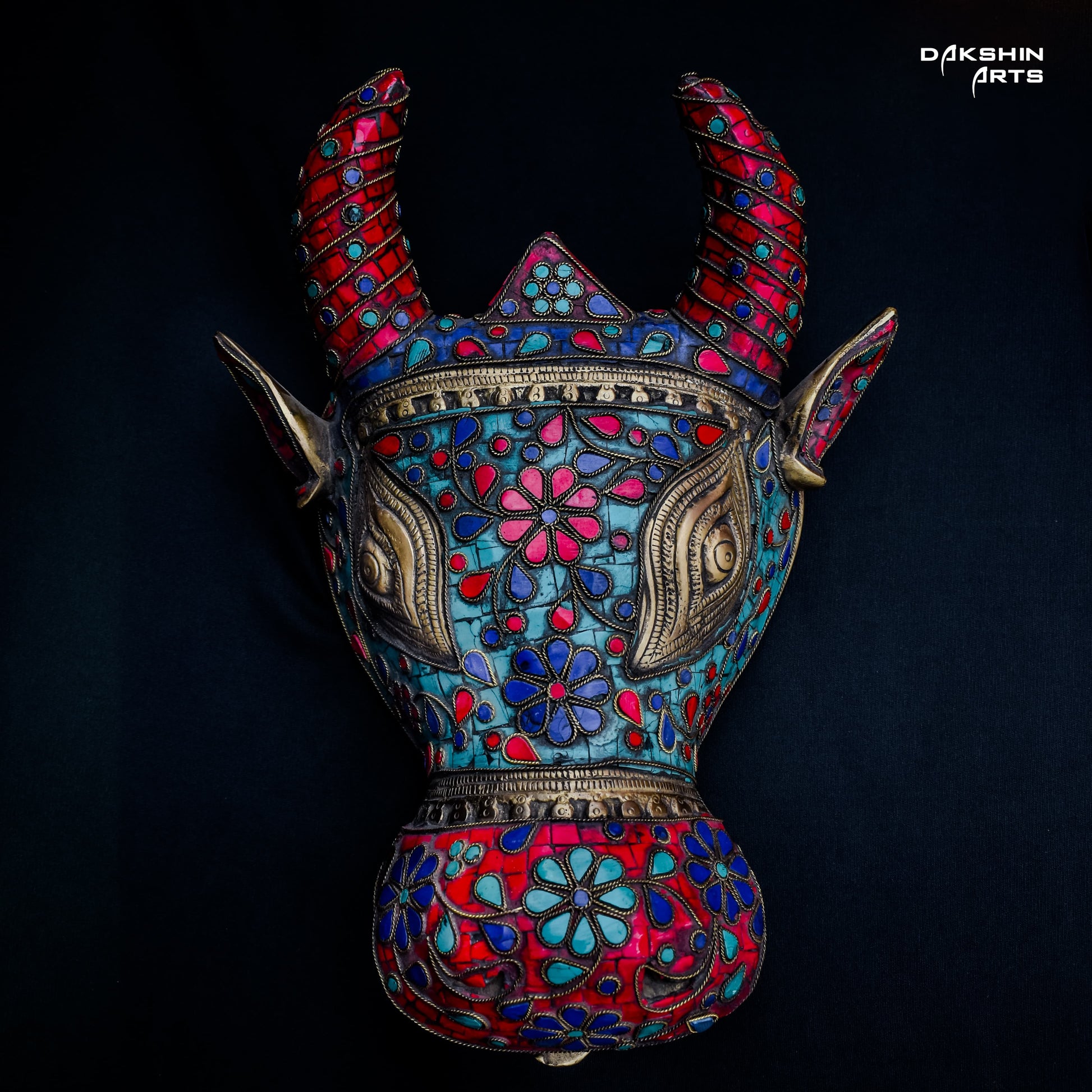 Colorful decorative brass Nandi mask with intricate floral patterns in red, blue, and turquoise on a black background- Dakshin Arts