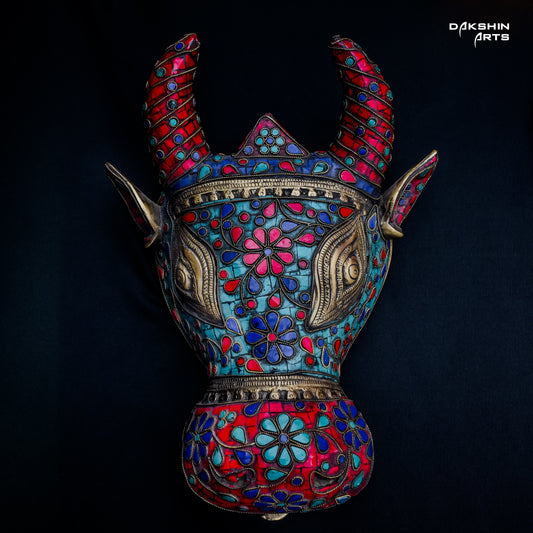 Colorful decorative brass Nandi mask with intricate floral patterns in red, blue, and turquoise on a black background- Dakshin Arts