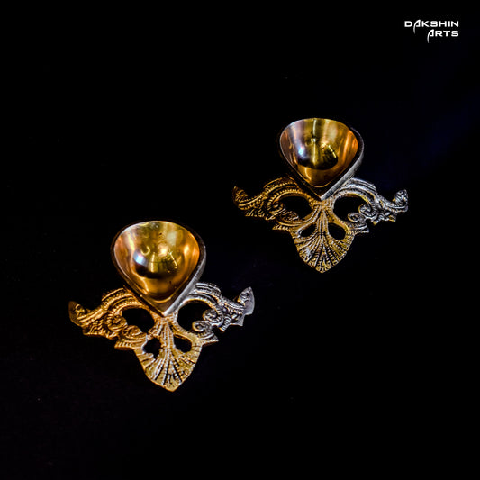 Two ornate, gold-tone, brass diyas on a black background with intricate floral patterns- Dakshin Arts
