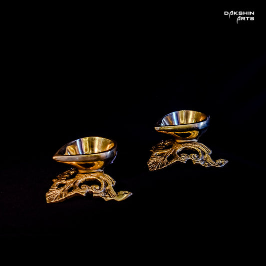 Two ornate, gold-tone, brass diyas on a black background with intricate floral patterns- Dakshin Arts