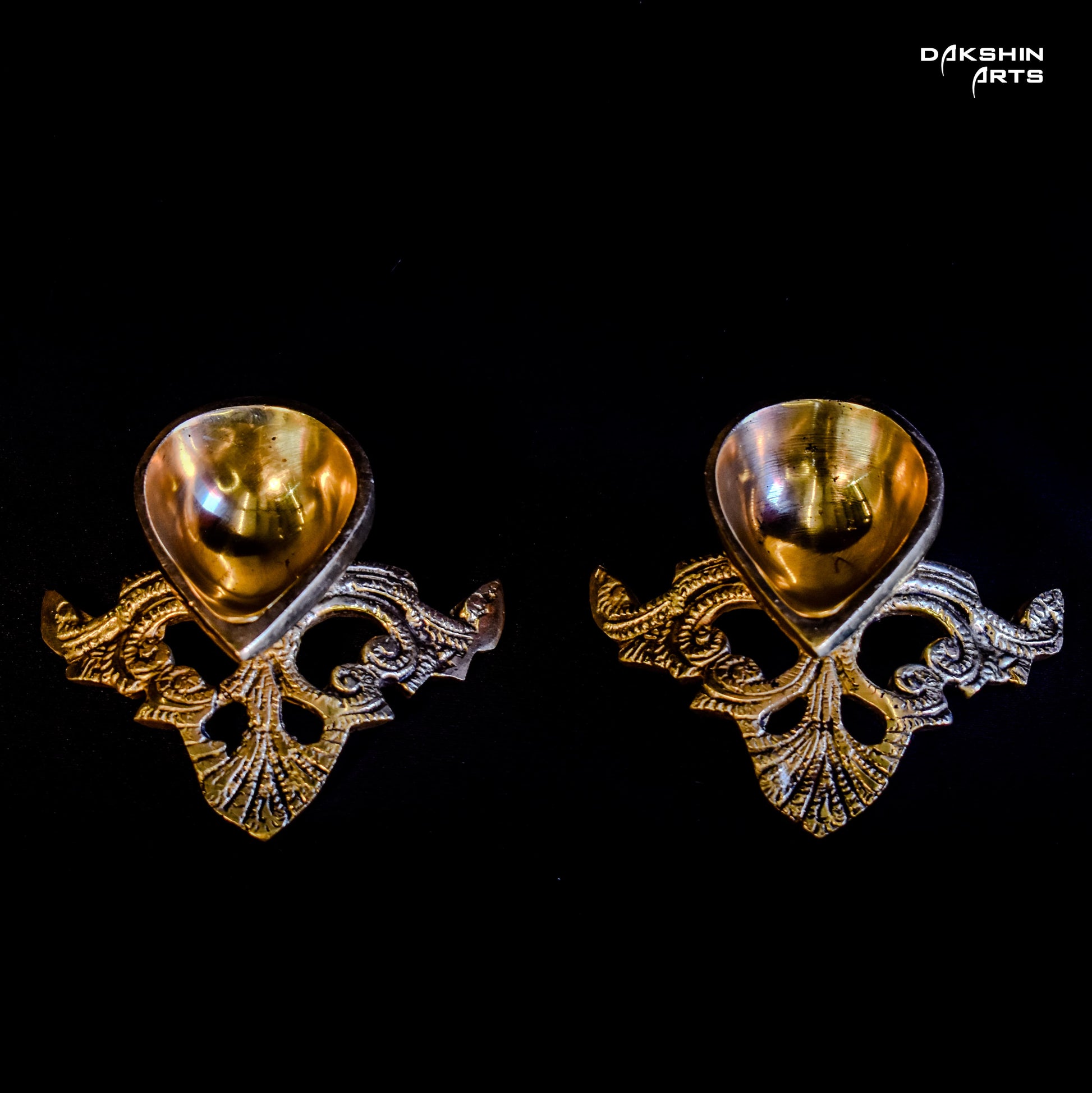 Two ornate, gold-tone, brass diyas on a black background with intricate floral patterns- Dakshin Arts