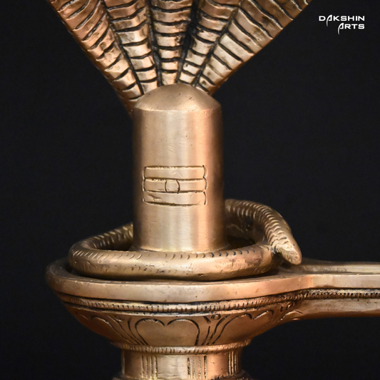 Brass sculpture of a Shivling with a five-headed cobra hood, set on a wooden surface against a black background- Dakshin Arts