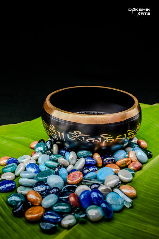 A black singing bowl with golden script, rests on vibrant green banana leaves with pebbles around - Dakshin Arts