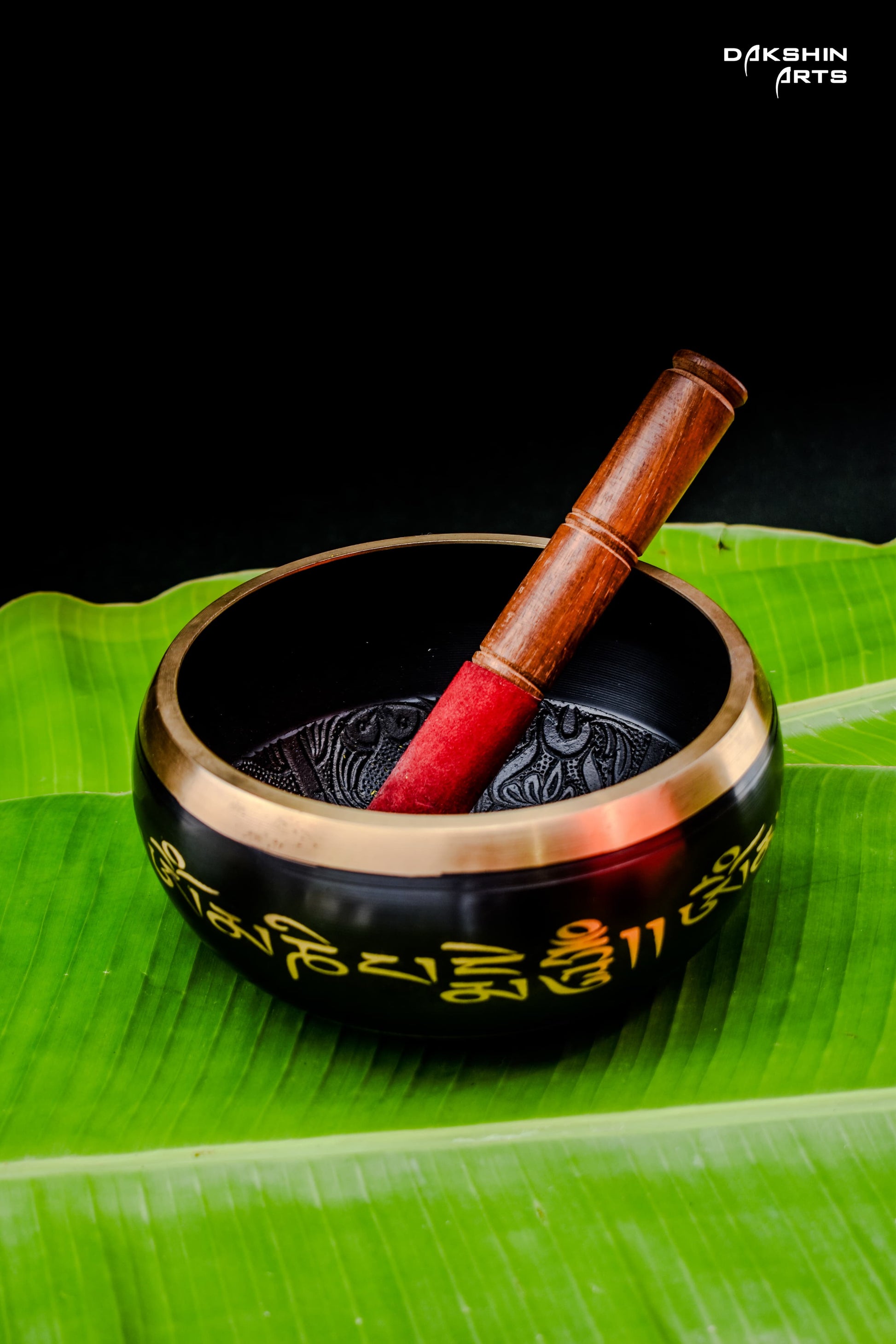 A black singing bowl with golden script with a wooden mallet, rests on green banana leaves- Dakshin Arts