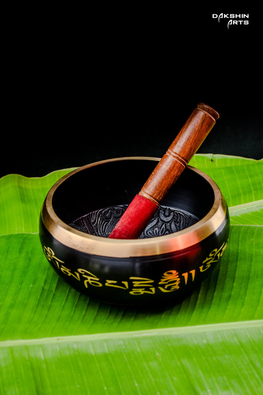 A black singing bowl with golden script with a wooden mallet, rests on green banana leaves- Dakshin Arts