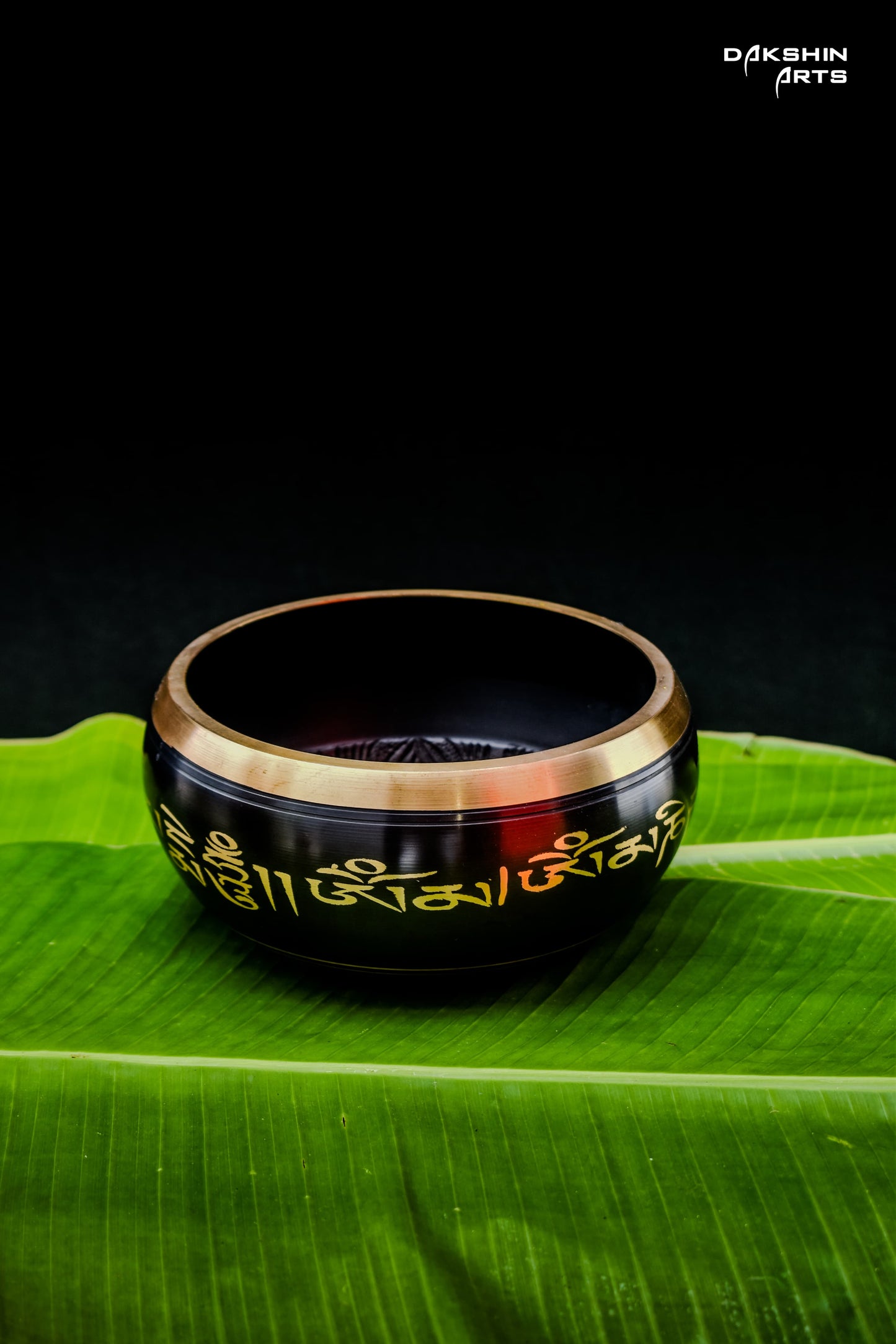 A black singing bowl with golden script, rests on green banana leaves- Dakshin Arts