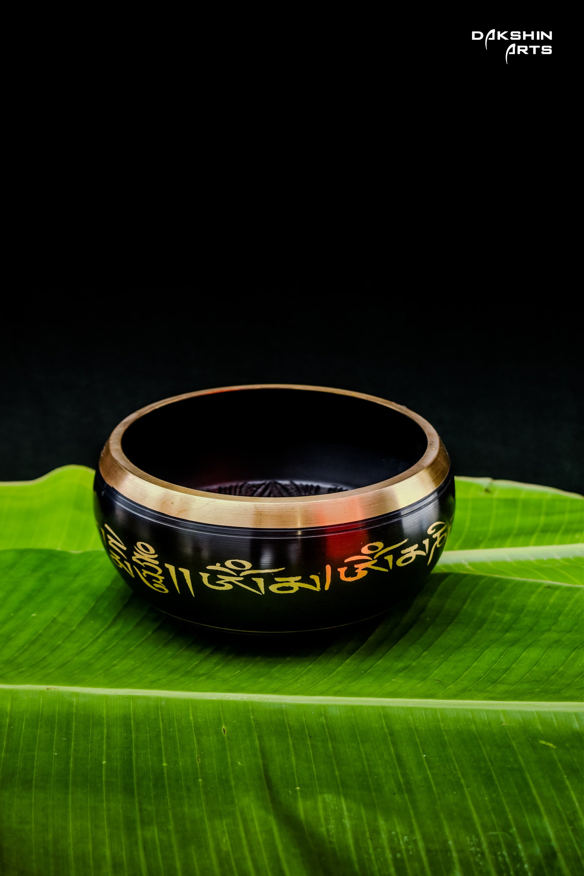 A black singing bowl with golden script, rests on green banana leaves- Dakshin Arts