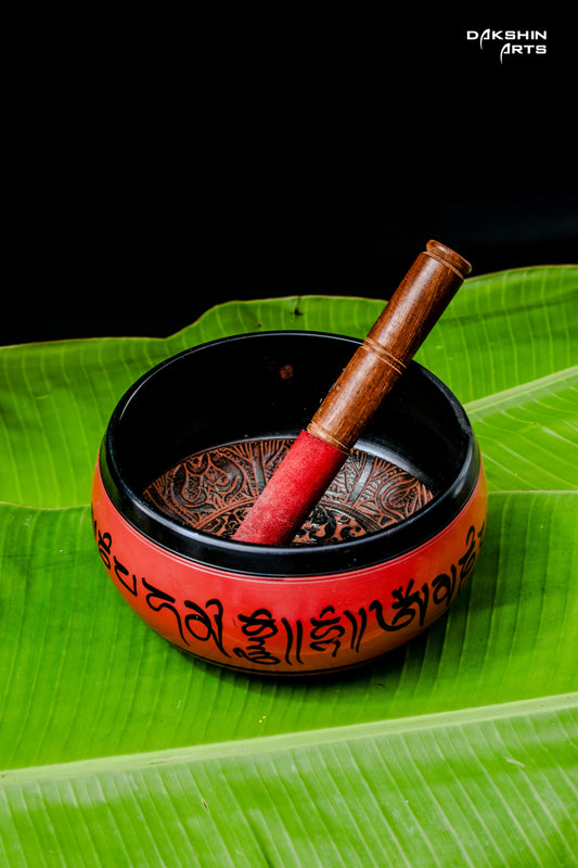 A red singing bowl with intricate patterns sits on vibrant green banana leaves. A wooden mallet rests inside- Dakshin Arts
