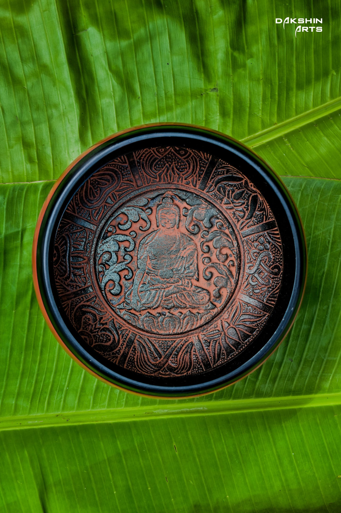 A red singing bowl with intricate patterns sits on vibrant green banana leaves - Dakshin Arts