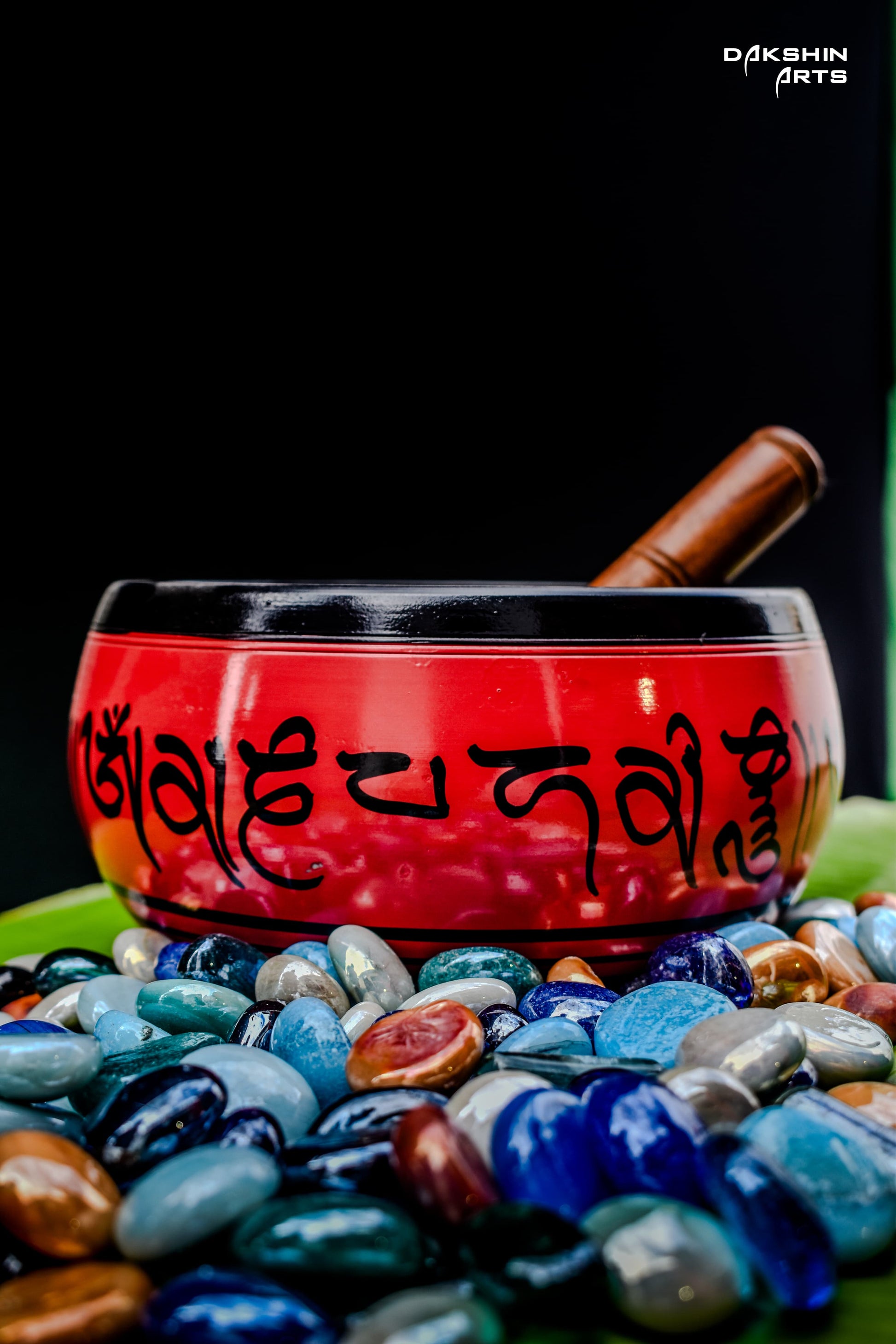 A red singing bowl with intricate patterns sits on green banana leaves surrounded by pebbles. A wooden mallet rests inside- Dakshin Arts