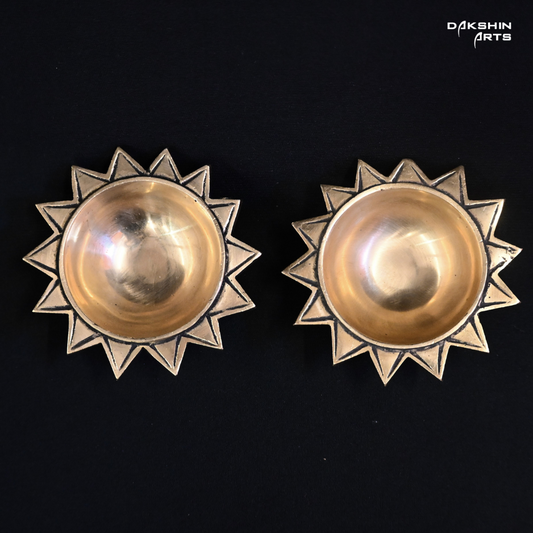 Two brass, sun-shaped diyas with star-like edges on a black background- Dakshin Arts