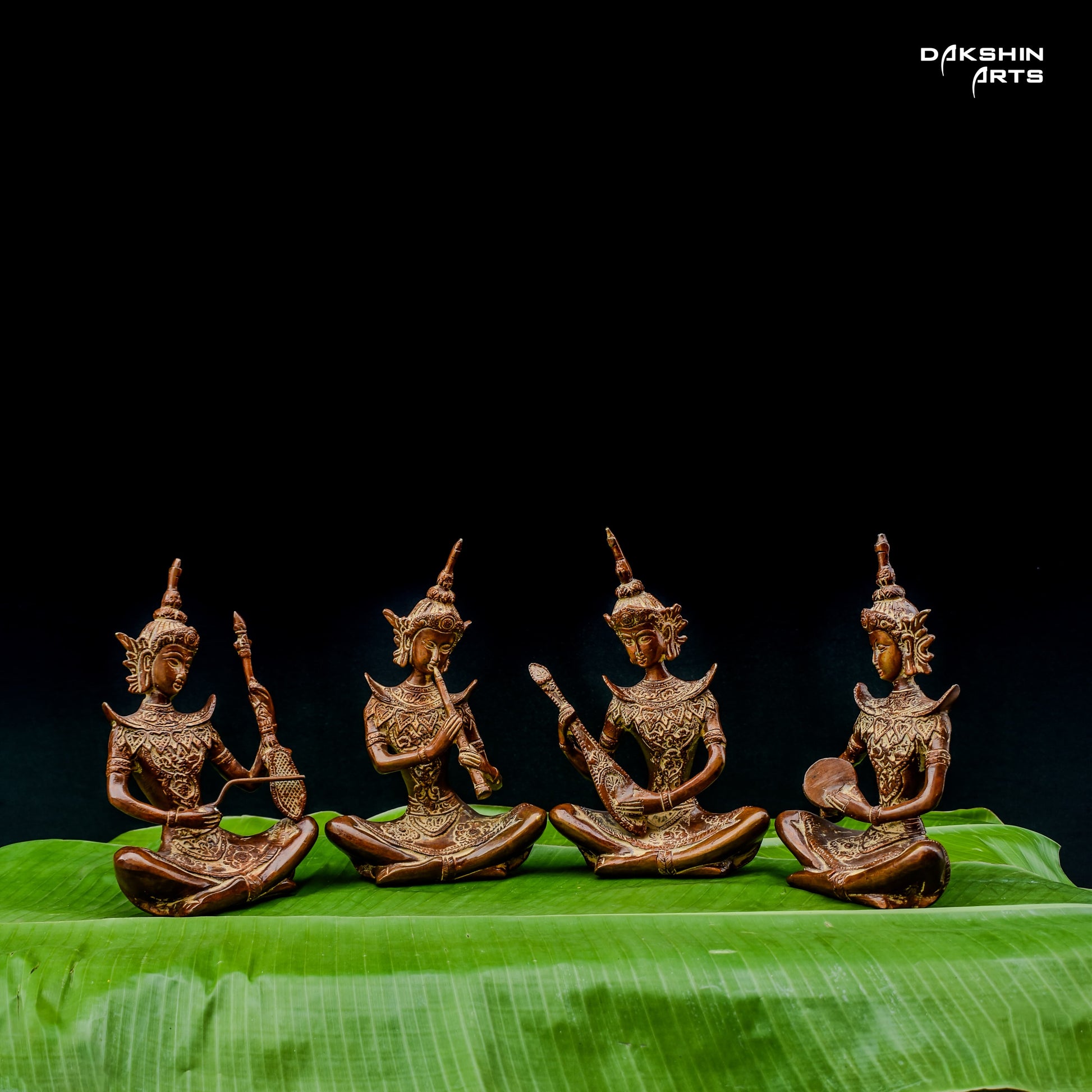 Four intricately carved brass statues of musicians are seated on vibrant green leaves against a black background- Dakshin Arts