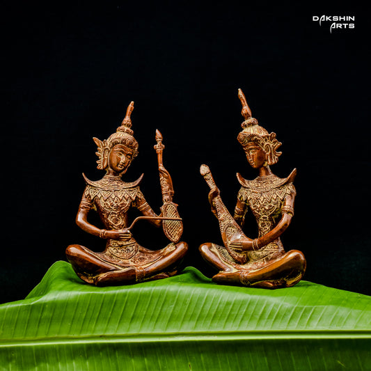 Two intricately carved brass statues of musicians are seated on vibrant green leaves against a black background- Dakshin Arts