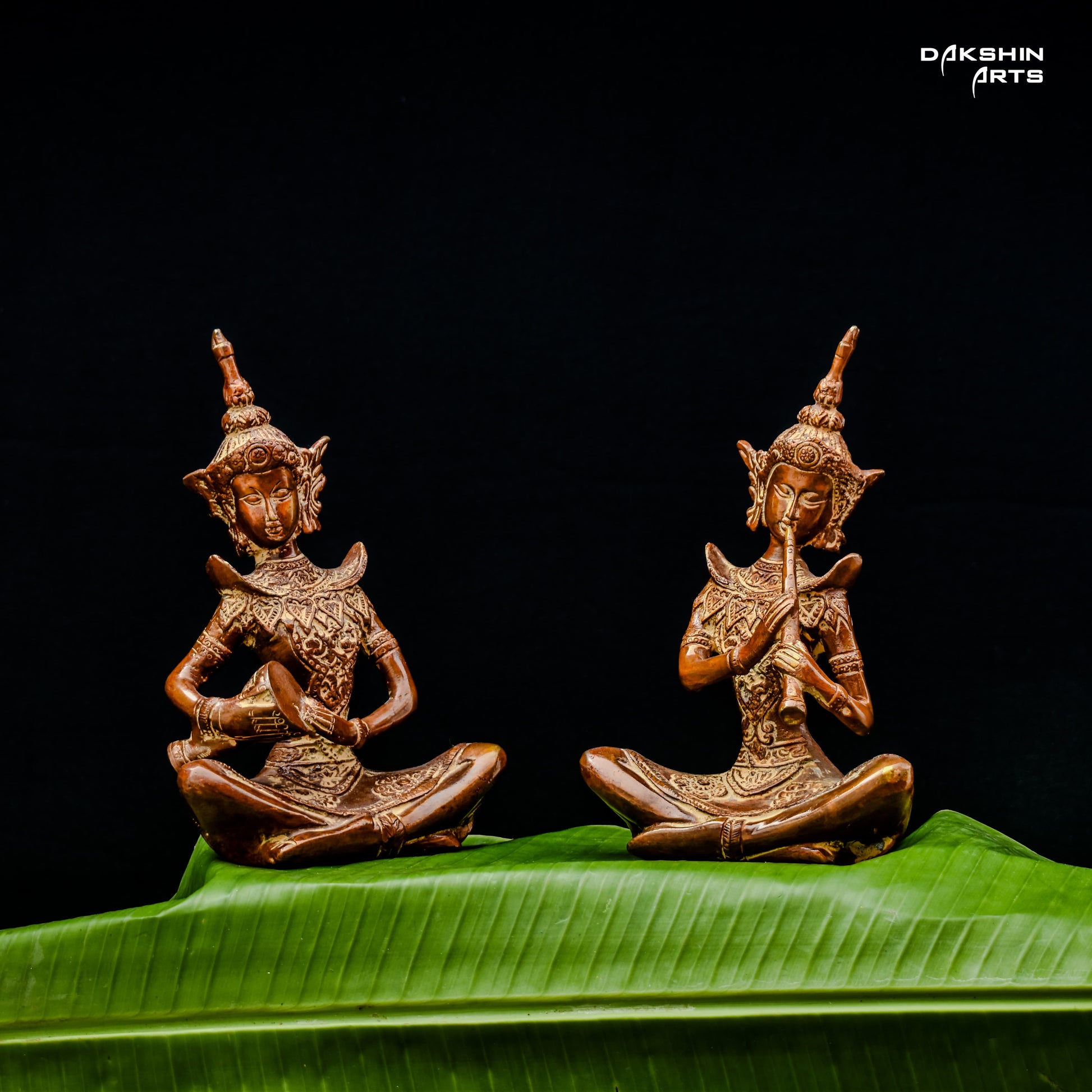 Two intricately carved brass statues of musicians are seated on vibrant green leaves against a black background- Dakshin Arts