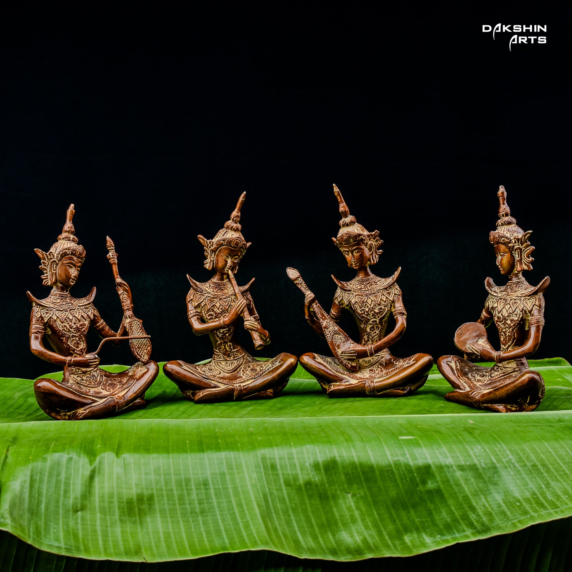 Four intricately carved brass statues of musicians are seated on vibrant green leaves against a black background- Dakshin Arts