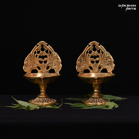 Two ornate brass diya lamps on a black background with 'Dakshin Arts' branding.