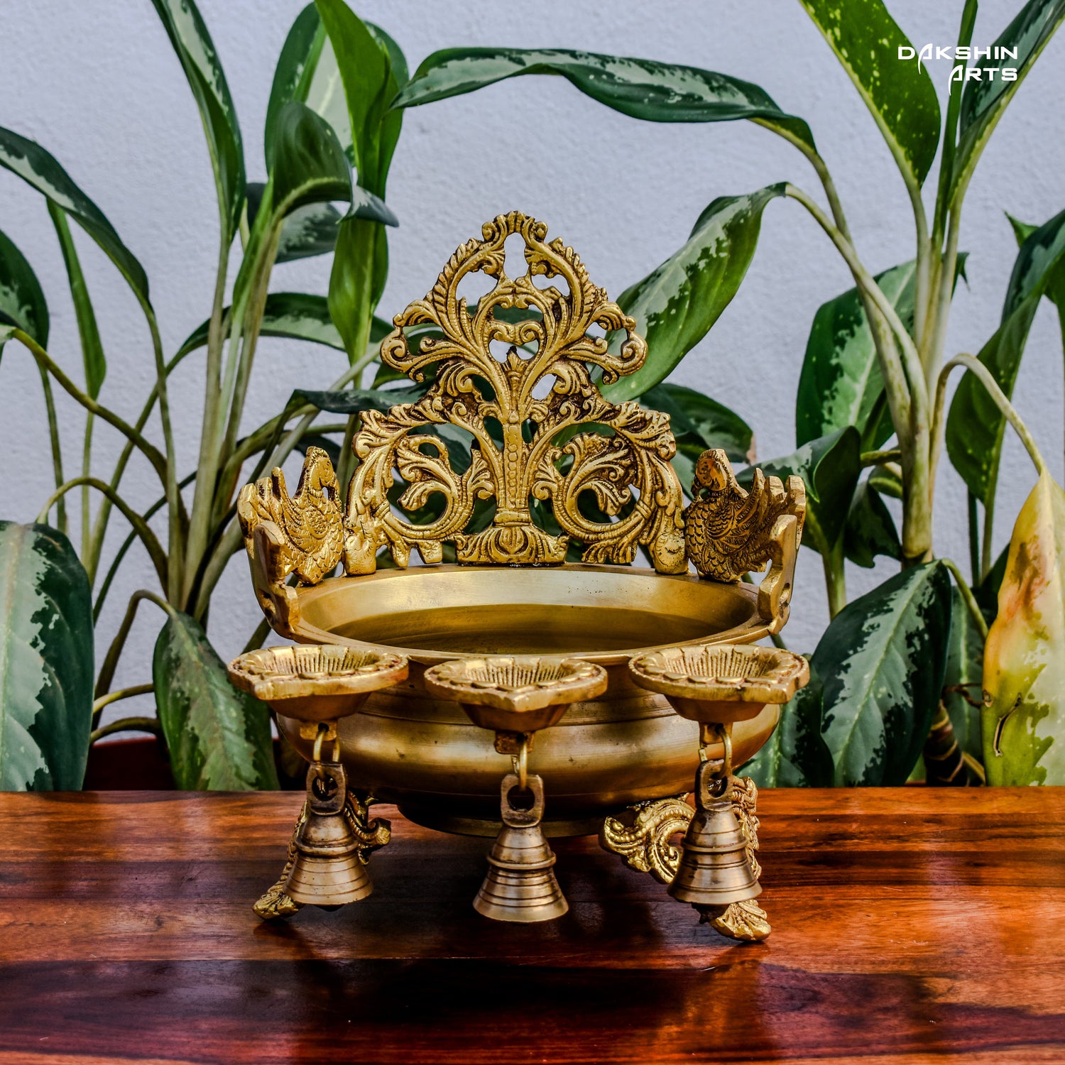 Ornate golden urli with oil lamps with intricate floral and peacock designs, set on a wooden table with leaves in the background- Dakshin Arts