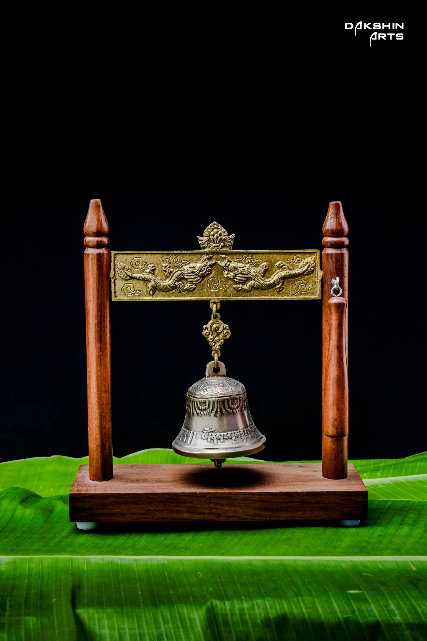 An ornate brass bell hangs in a wooden frame with intricate designs, displayed on vibrant green banana leaves - Dakshin Arts