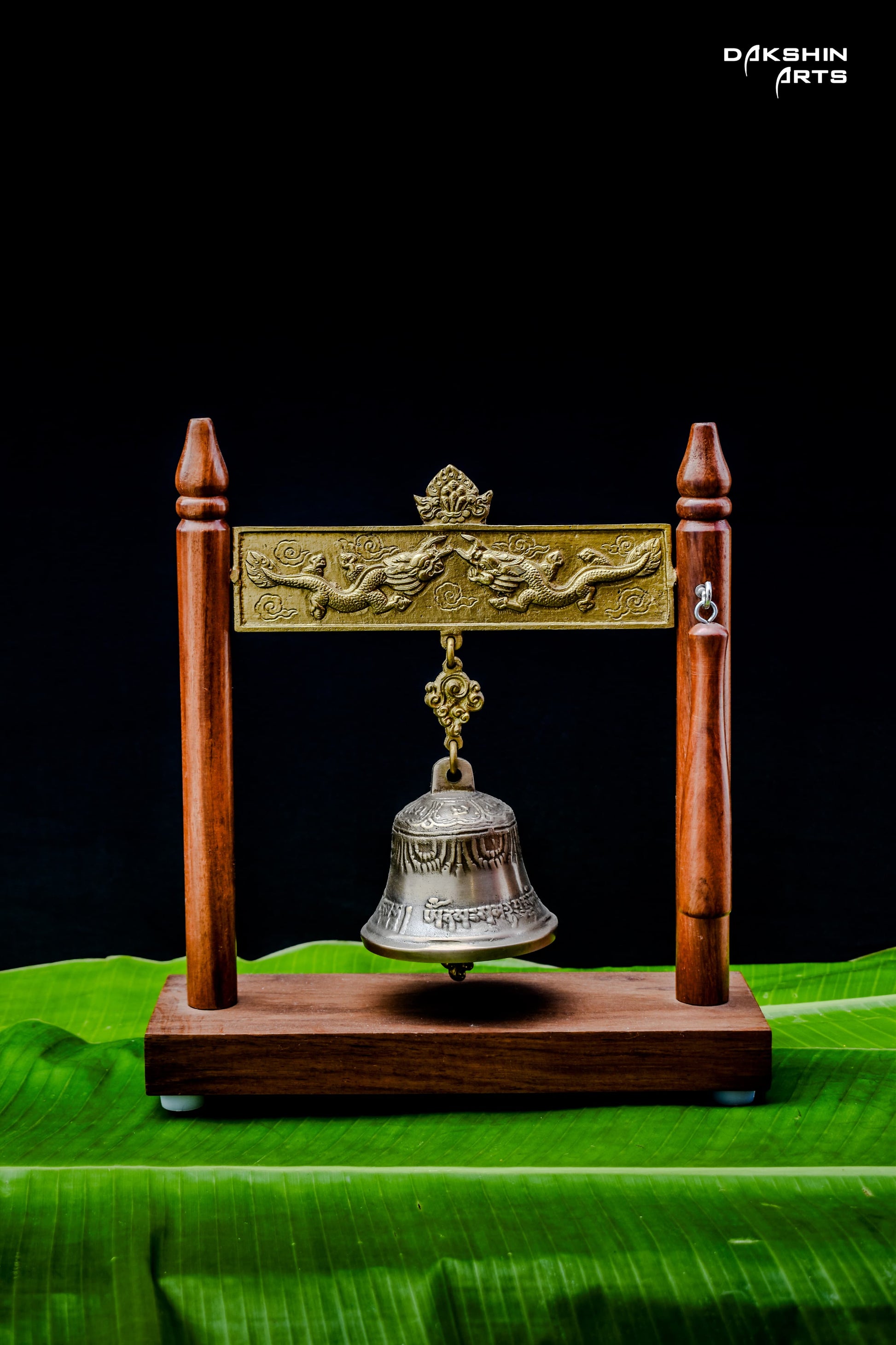 An ornate brass bell hangs in a wooden frame with intricate designs, displayed on vibrant green banana leaves - Dakshin Arts