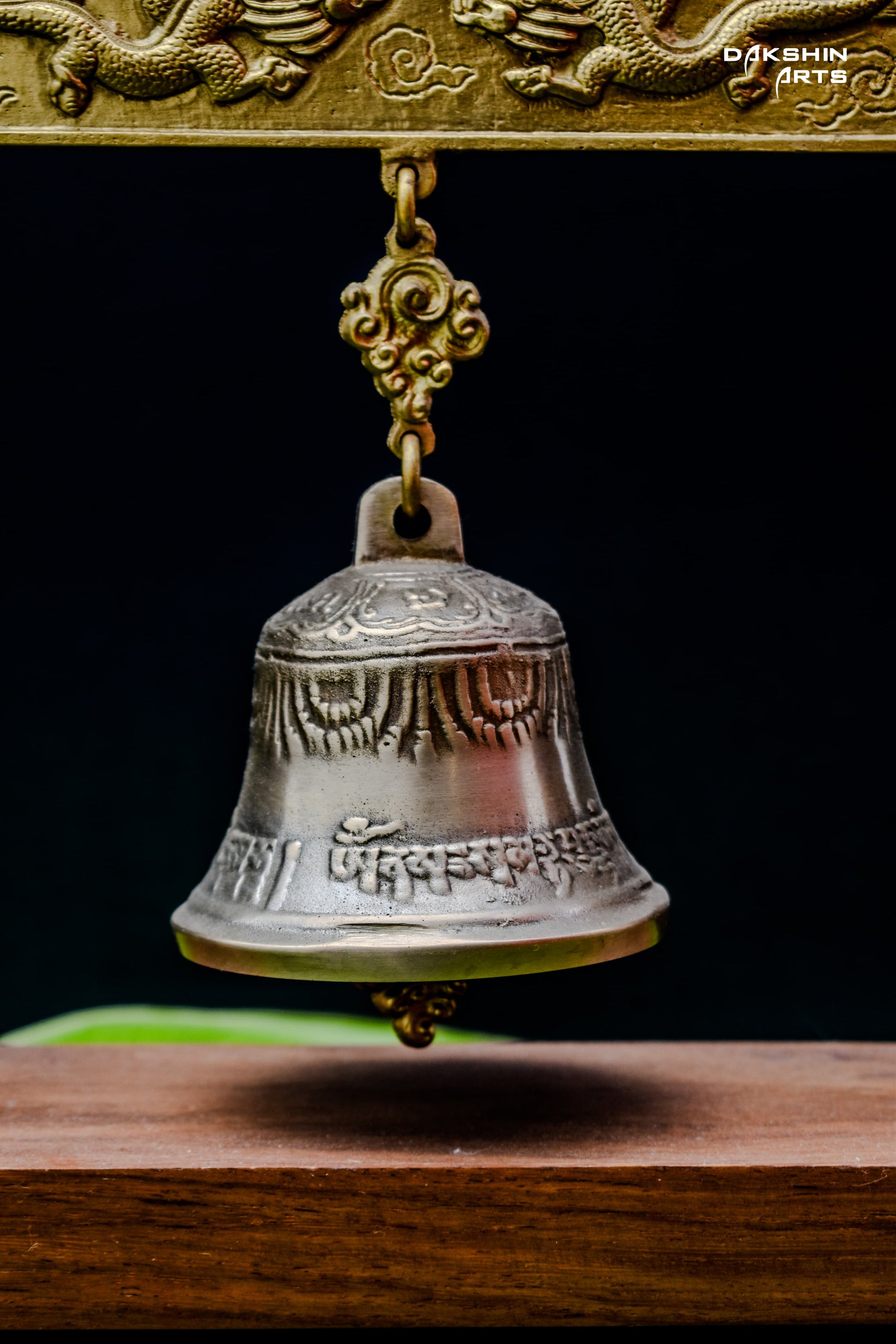 An ornate brass bell hangs in a wooden frame with intricate designs, displayed on vibrant green banana leaves - Dakshin Arts