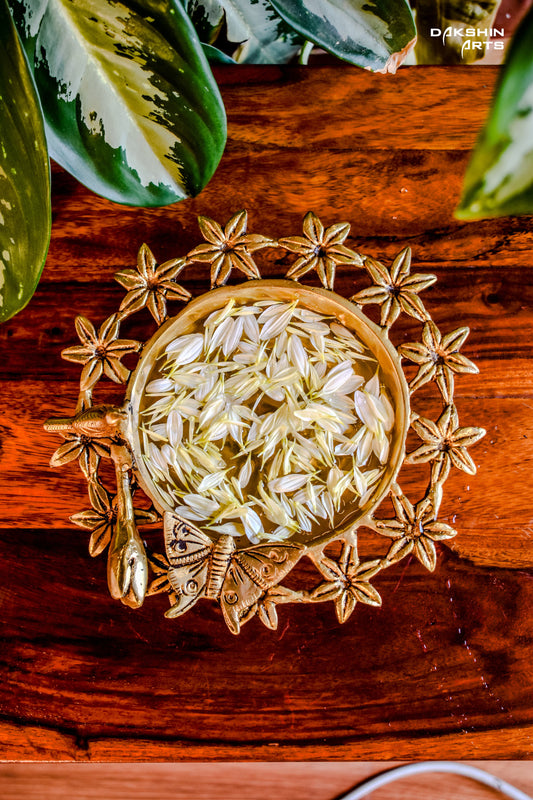 Brass Urli with floral motifs and a butterfly on wooden table filled with white petals. Green leaves peek from the top corner- Dakshin Arts