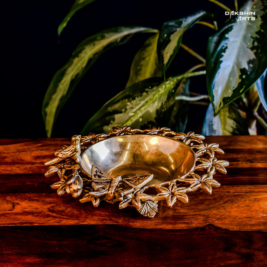 Brass Urli with floral motifs and a butterfly on wooden table. Green leaves peek from the top corner- Dakshin Arts